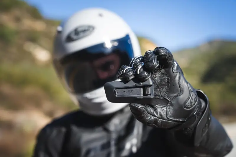 Rider-Showcasing-the-Cardo-Packtalk-Pro A close-up shot of a motorcyclist's gloved hand holding a black cardo packtalk pro communication system, with their white helmet visible in the blurred background..