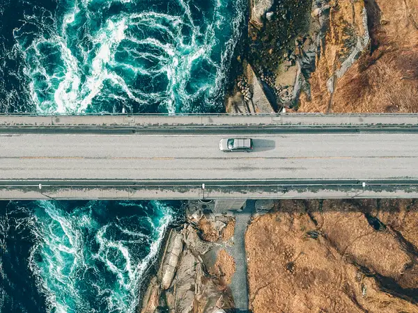 Highway-Area Top-down aerial view of a silver car driving across a bridge spanning turbulent blue water between rocky shores.