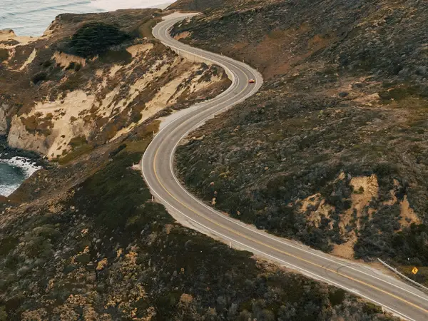 Mountain-Area Aerial view of a winding road snaking along rugged coastal cliffs next to the ocean.