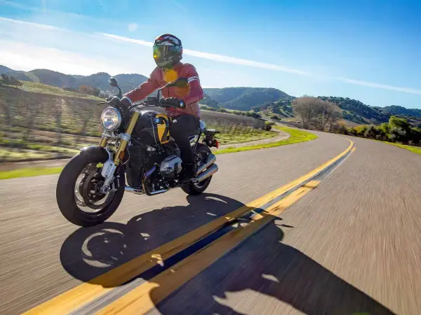Helmet-Speaker-Rider A motorcyclist wearing a full-face helmet, possibly enjoying audio from a helmet speaker, rides a black and yellow motorcycle down a sun-drenched, winding paved road through rolling hills.
